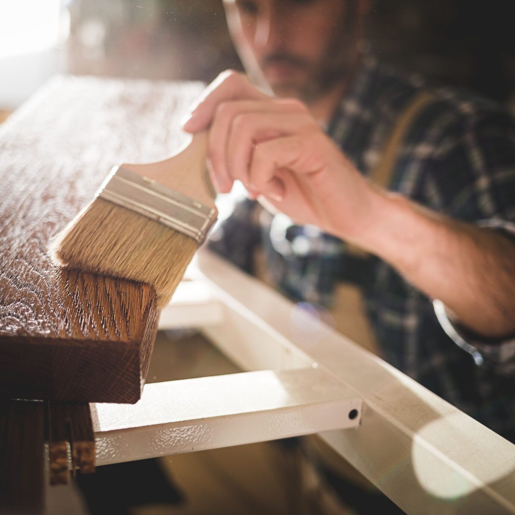 Close up of paintbrush applies paint or varnish on wooden board in carpentry workshop